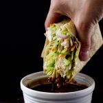 A black background, with a hand holding a tortilla wrap containing veggie slaw and chicken being dipped in a dark brown sauce in a small white bowl.