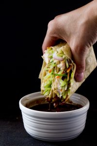 A black background, with a hand holding a tortilla wrap containing veggie slaw and chicken being dipped in a dark brown sauce in a small white bowl.