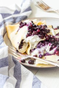 3 crepes folded in triangled are stuffed with a white filling and topped with lots of blueberries. A fork sits on the plate and a blue and white striped towel is crumpled on the left side.