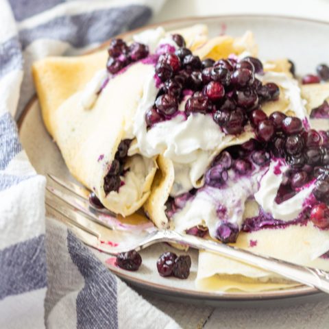 3 crepes folded in triangled are stuffed with a white filling and topped with lots of blueberries. A fork sits on the plate and a blue and white striped towel is crumpled on the left side.