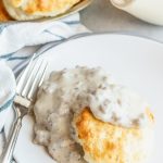 A biscuit sits on a round white plate topped with a gray sausage gravy. A fork sits angled on the side of a plate and a soft white and blue striped towel is underneath. A bowl of biscuits and a gravy boat are behind the plate.