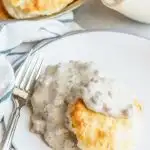 A biscuit sits on a round white plate topped with a gray sausage gravy. A fork sits angled on the side of a plate and a soft white and blue striped towel is underneath. A bowl of biscuits and a gravy boat are behind the plate.