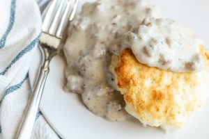 A biscuit sits on a round white plate topped with a gray sausage gravy. A fork sits angled on the side of a plate and a soft white and blue striped towel is underneath