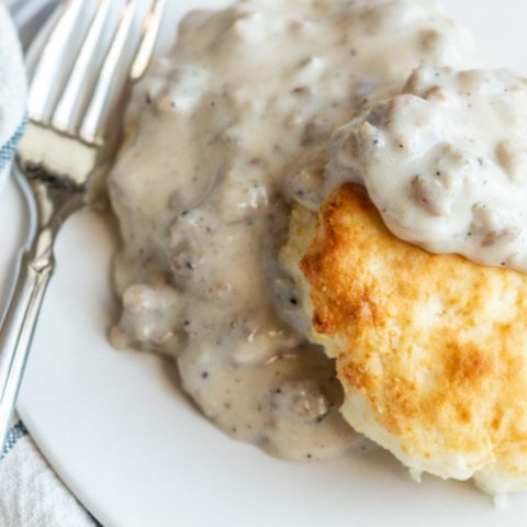 A biscuit sits on a round white plate topped with a gray sausage gravy. A fork sits angled on the side of a plate and a soft white and blue striped towel is underneath