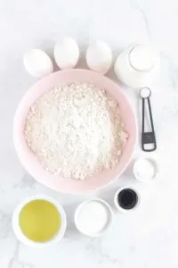 ingredients in bowls: 3 eggs, salt, baking powder/soda, vanilla, sugar, oil, milk, and a large pink bowl of flour
