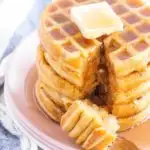 stack of gluten free waffles on a pink plate. Topped with a pat of butter and drizzled with maple syrup. A bite on a fork is sitting on the plate.