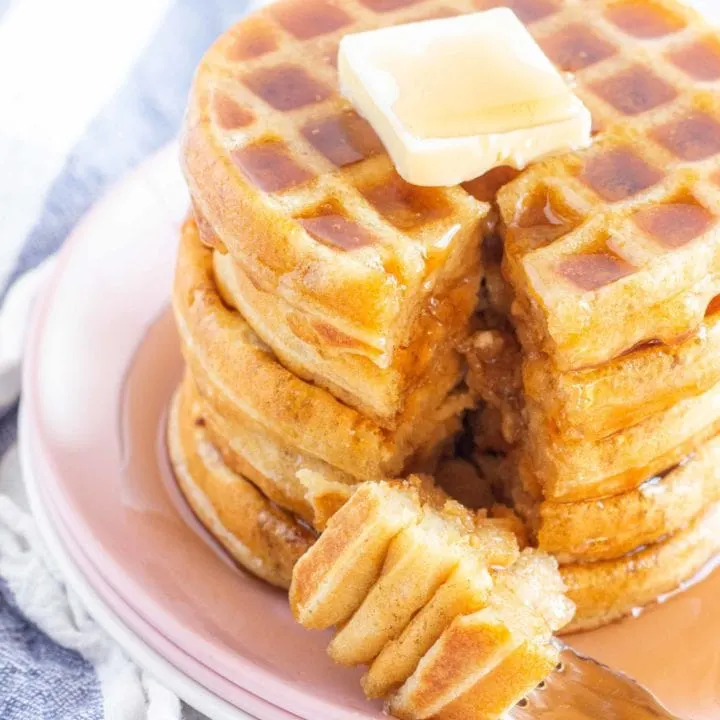 stack of gluten free waffles on a pink plate. Topped with a pat of butter and drizzled with maple syrup. A bite on a fork is sitting on the plate.