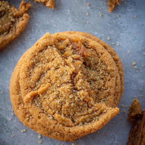 A stack of pumpkin cookies with half a cookie on top and crumbs surrounding it. Cookies are on a dark blue and white marbled backdrop and everything is sprinkled with coarse sugar.