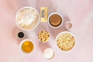 Ingredients are set out in white ceramic bowls on a pink and white marbled surface. In the center is a medium bowl roughly chopped macadamia nuts. Clockwise from top left: large bowl of gluten free flour, a stick of butter, medium bowl of dark brown sugar, small bowl of sea salt and baking soda, large bowl of white chocolate chips, medium bowl of white sugar, medium bowl with 1 whole egg plus 1 egg yolk, and small bowl with dark vanilla extract