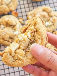 a hand holds a gluten free white chocolate macadamia nut cookie which has been broken in half. The halves are stacked, bottoms together in the center. The inside looks chewy and the outside edges are a golden brown. The top is crinkly and studded with macadamia nut chunks and white chocolate chips. There is a sprinkling of a flaky salt. In the background you can see more cookies on a black cooling rack.