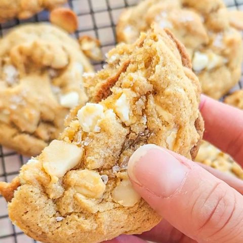 a hand holds a gluten free white chocolate macadamia nut cookie which has been broken in half. The halves are stacked, bottoms together in the center. The inside looks chewy and the outside edges are a golden brown. The top is crinkly and studded with macadamia nut chunks and white chocolate chips. There is a sprinkling of a flaky salt. In the background you can see more cookies on a black cooling rack.