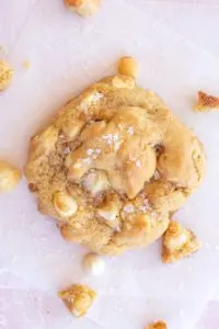 A single white chocolate macadamia nut cookie sits on 2 layers of offset parchment paper on top of a pink and white marbled surface. there are chunks of macadamia nuts, cookie crumbs, and a white chocolate chip scattered around the cookie. The cookie is thick with a wrinkled top and golden brown highlights. It is sprinkled with flaky salt.