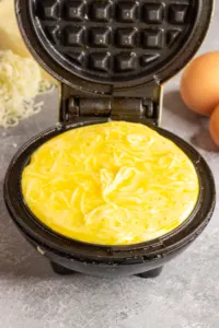a mini waffle iron is open and filled with a mixture of eggs and cheese. In the background you can see 2 brown eggs and a pile of finely grated white cheese