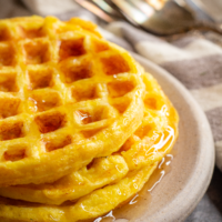 a stack of 3 round waffles on a gray stone plate. They are topped with syrup with is running down the sides and pooling on the plate. A white and gray striped linen is in the background along with a stack of silver forks