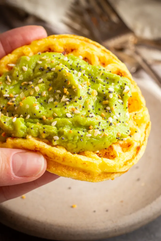 a hand holds a small round chaffle. It is topped with mashed avocado and sprinkled with everything bagel seasoning