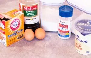 From left to right: a box of baking soda, bottle of Mexican vanilla, 2 brown eggs, a canister of gluten free flour, Redmond Real Salt, and a canister of sugar, and a tub of Tillamook sour cream