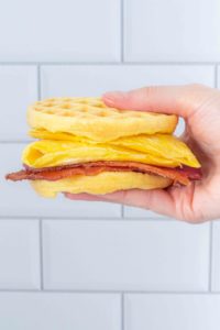 a hand holds a breakfast sandwich made with two wonder bread chaffles, strips of bacon, and an omelet in front of a white tile background