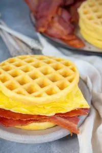 a chaffle breakfast sandwich made with bacon and eggs sits on a stone plate on top of a gray marbled background. Next to the plate is a white linen napkin and behind is a pile of forks and a plate with stacks of more keto chaffles and bacon strips.