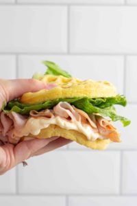 a hand holds a sandwich made with folded lunch meat, large pieces of lettuce, and mayonnaise which is oozing over the side. the wonder bread chaffles are soft and bendy