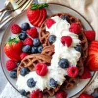 a plate of gluten free chocolate waffles slightly overlapping on a plate. Covered in whipped cream, strawberries, raspberries, and blueberries