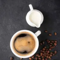 A mug off coffee sits on a black slate background. Next to it is a white pitcher full of creamer and a scattering of coffee beans