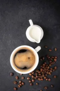 A mug off coffee sits on a black slate background. Next to it is a white pitcher full of creamer and a scattering of coffee beans