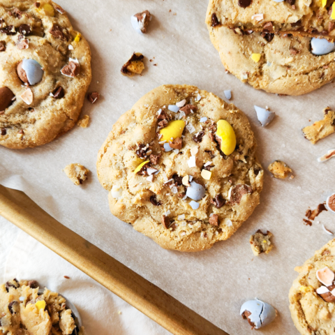 Gluten free cadbury egg cookies sit on a baking sheet lined with parchment paper. Crumbs and pieces of Cadbury mini eggs are scattered around the cookies, and a cookie cut in half sits on top of one of the cookies so you can see the texture. It is chewy and doughy inside. Next to the baking sheet is a cookie scoop filled with cookie dough.