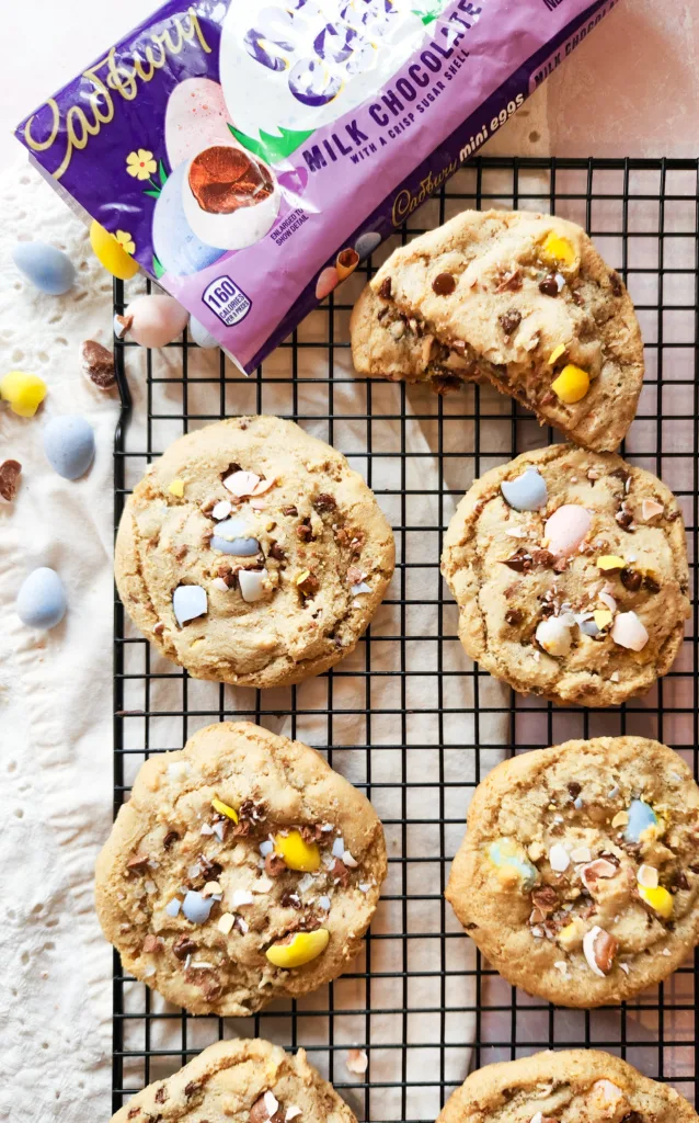 cookies sit on a cooling rack. They are studded with crushed Cadbury mini eggs and mini chocolate chips. A bag of Cadbury mini eggs is on the top left corner and mini eggs are spilling out of it.