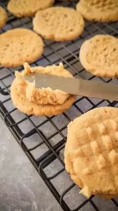 a knife is spreading peanut butter frosting on the smooth side of a peanut butter cookie.