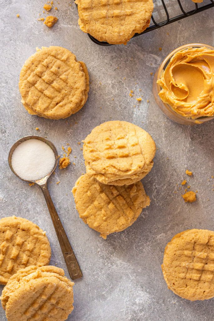 gluten free nutter butter sandwich cookies and peanut butter cookies are scattered on a gray marbled backdrop. a glass bowl of creamy peanut butter sits in the top right corner, and a spoon of white sugar is laid on the left side.