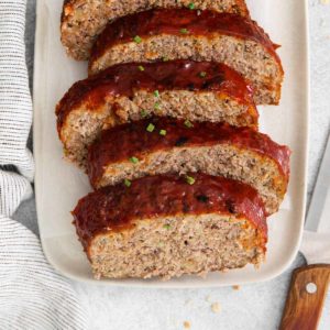 Slices of meatloaf are on a white serving tray. On one side of the tray is a knife, and on the other is a soft linen.