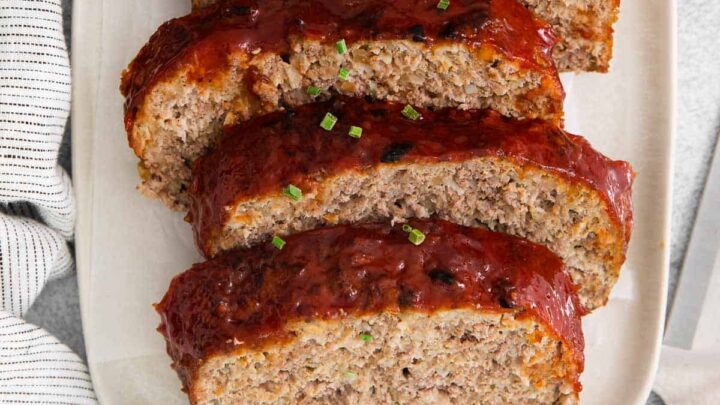 Slices of meatloaf are on a white serving tray. On one side of the tray is a knife, and on the other is a soft linen.