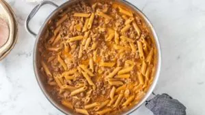 a skillet sits on a white background. Inside the skillet is a pasta dish with penne pasta and ground beef in a tomato sauce.
