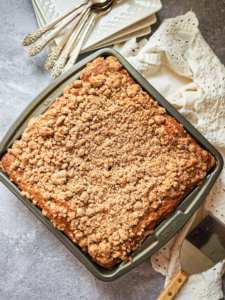 a coffee cake with lots of crumb topping in a square pan sits on a gray marbled coutertop. Plates, a white lacey napkin, and silver forks are scattered nearby.