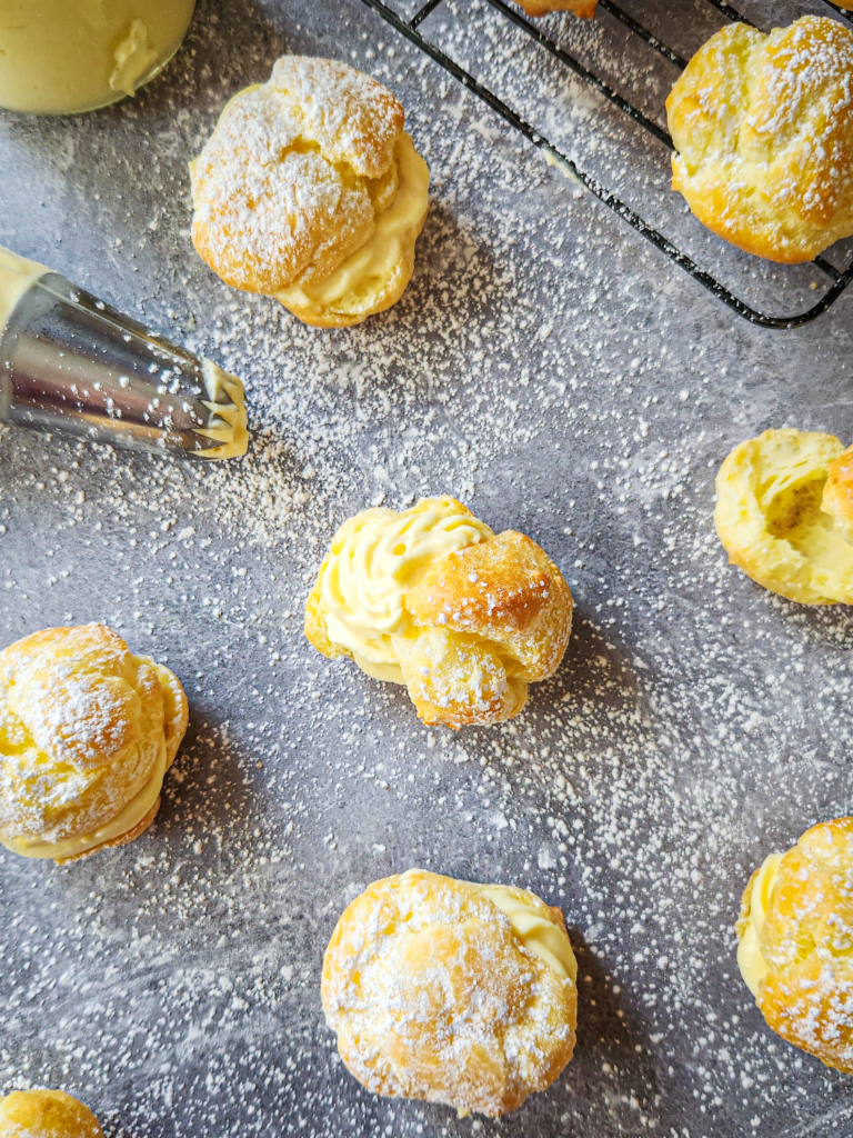 cream puffs are scattered on a gray marble background. Some are filled with vanilla pastry cream , some are cut open to reveal their hollow inside. Everything is dusted with powdered sugar and there is a pastry bag fitted with a decorating tip used for the vanilla pastry cream.
