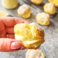 a hand holds a gluten-free cream puff. It is sliced in half and filled with a creamy vanilla filling. Empty and filled baked choux pastry sit on the counter and a cooling rack in the background.