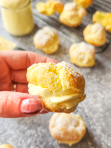 a hand holds a gluten-free cream puff. It is sliced in half and filled with a creamy vanilla filling. Empty and filled baked choux pastry sit on the counter and a cooling rack in the background.