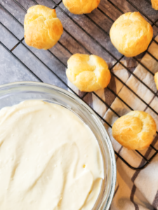 cream puff shells sit on a cooling rack over a gray marble background next to a bowl of vanilla pastry cream.