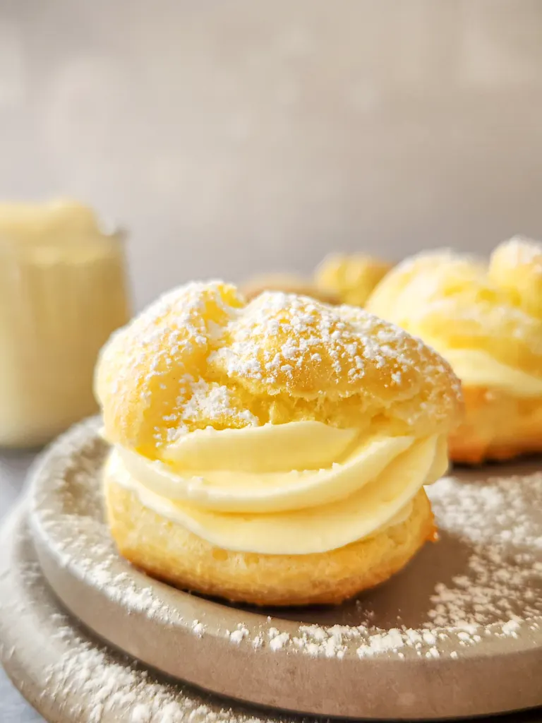 a cream puff filled with vanilla pastry cream sits on a gray stone plate. More cream purrs are in the background, and a glass jar is filled with vanilla pastry cream.