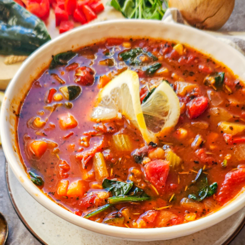 A white bowl is filled with vegetable soup and topped with a twisted lemon slice. An onion, parsley, and red and green peppers are in the background.