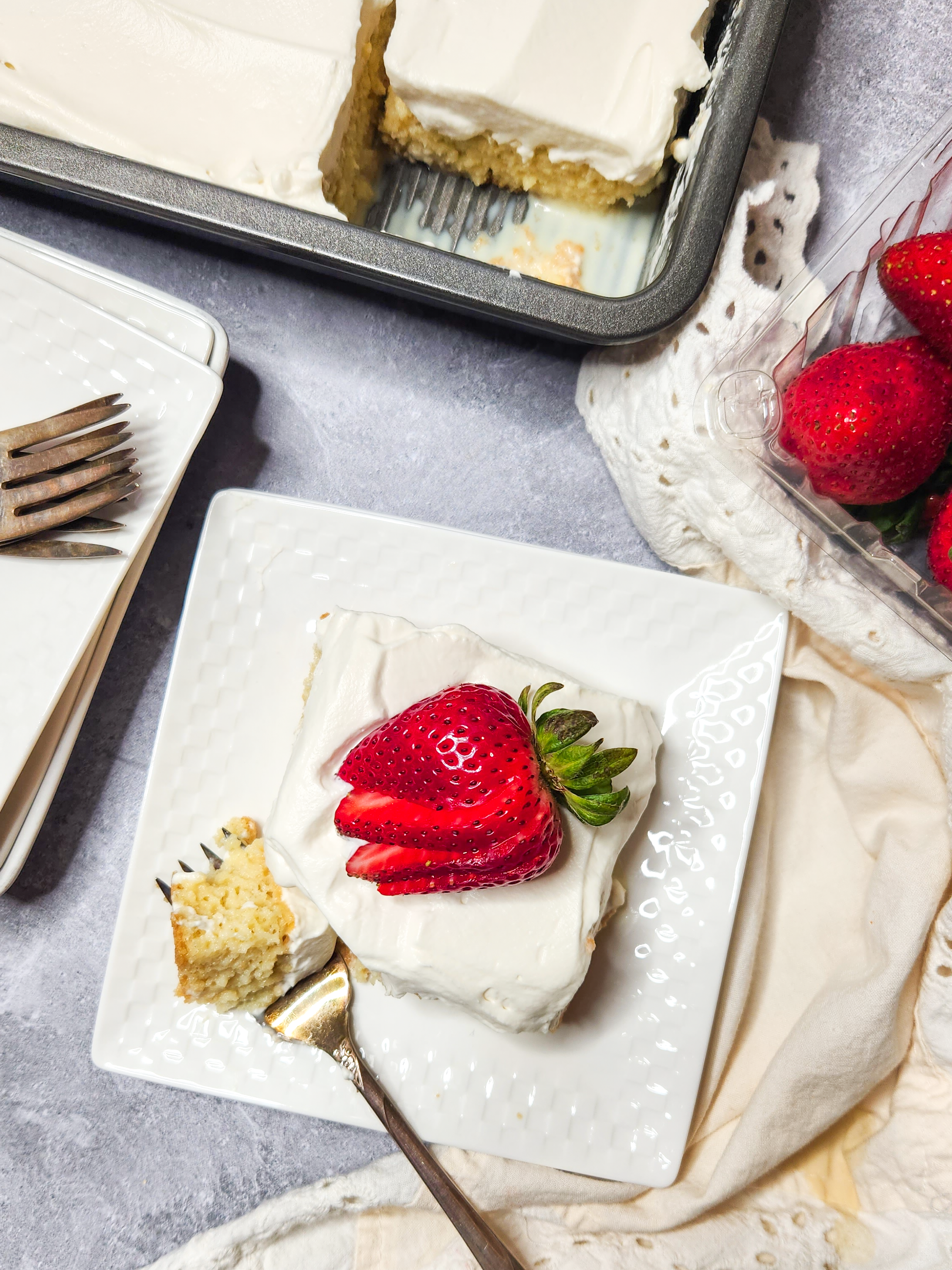 An overhead photo featuring a pan of gluten free tres leches with one slice out. There is a small puddle of milk in the bottom of the pan.  A slice of cake is beside the pan on a white ceramic plate.