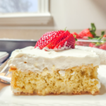 a slice of gluten free tres leches sits on a white ceramic plate. It is topped with a thick layer of whipped cream and sliced strawberries.