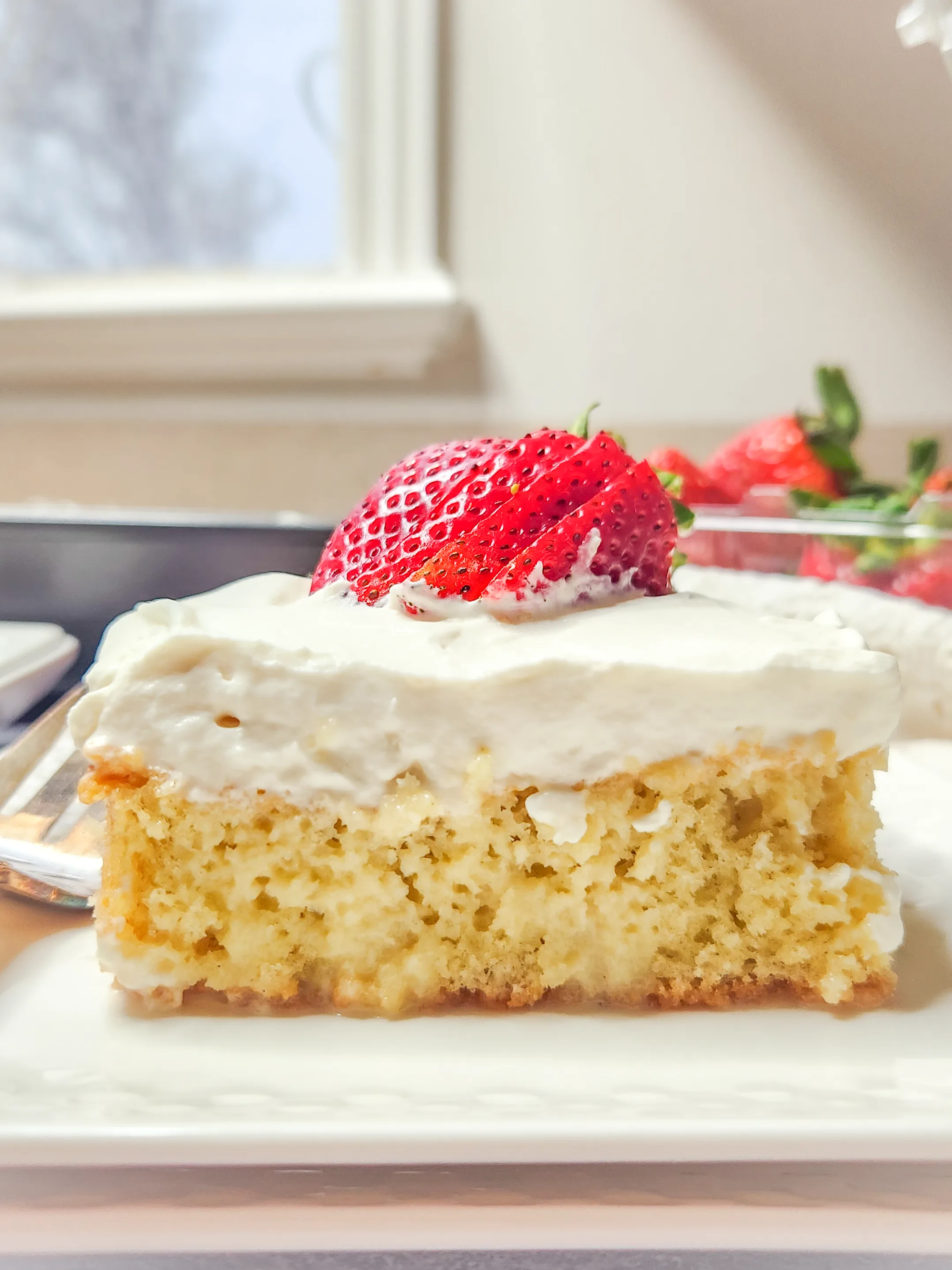 a slice of gluten free tres leches sits on a white ceramic plate. It is topped with a thick layer of whipped cream and sliced strawberries.