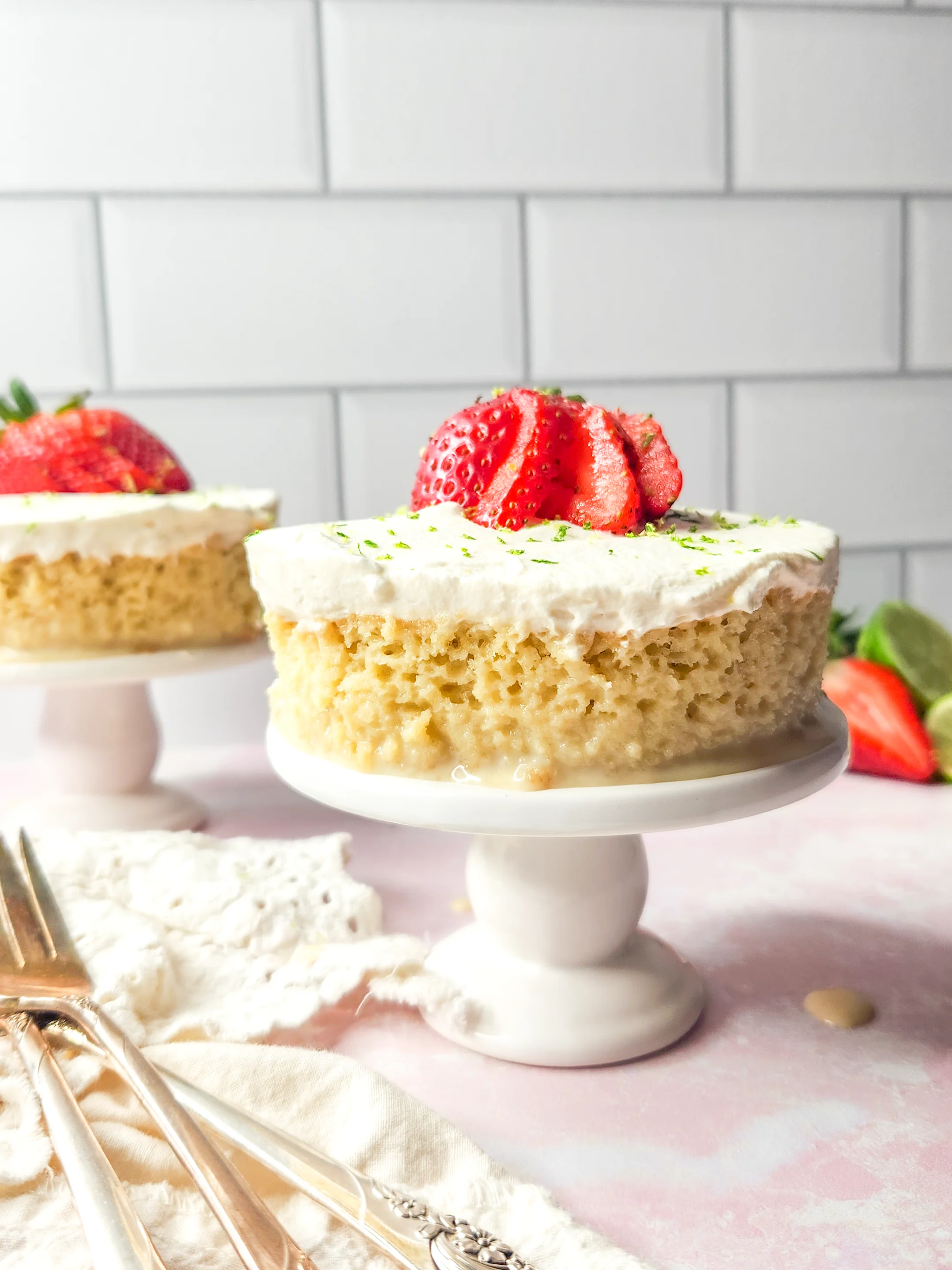 A round cut-out of gluten free tres leches cake sits on a white mini cake stand. It is frosted with whipped cream and topped with sliced strawberries and a sprinkling of lime zest. Another mini cake stand features the same in the background, and additional strawberries and limes can be seen in the background.