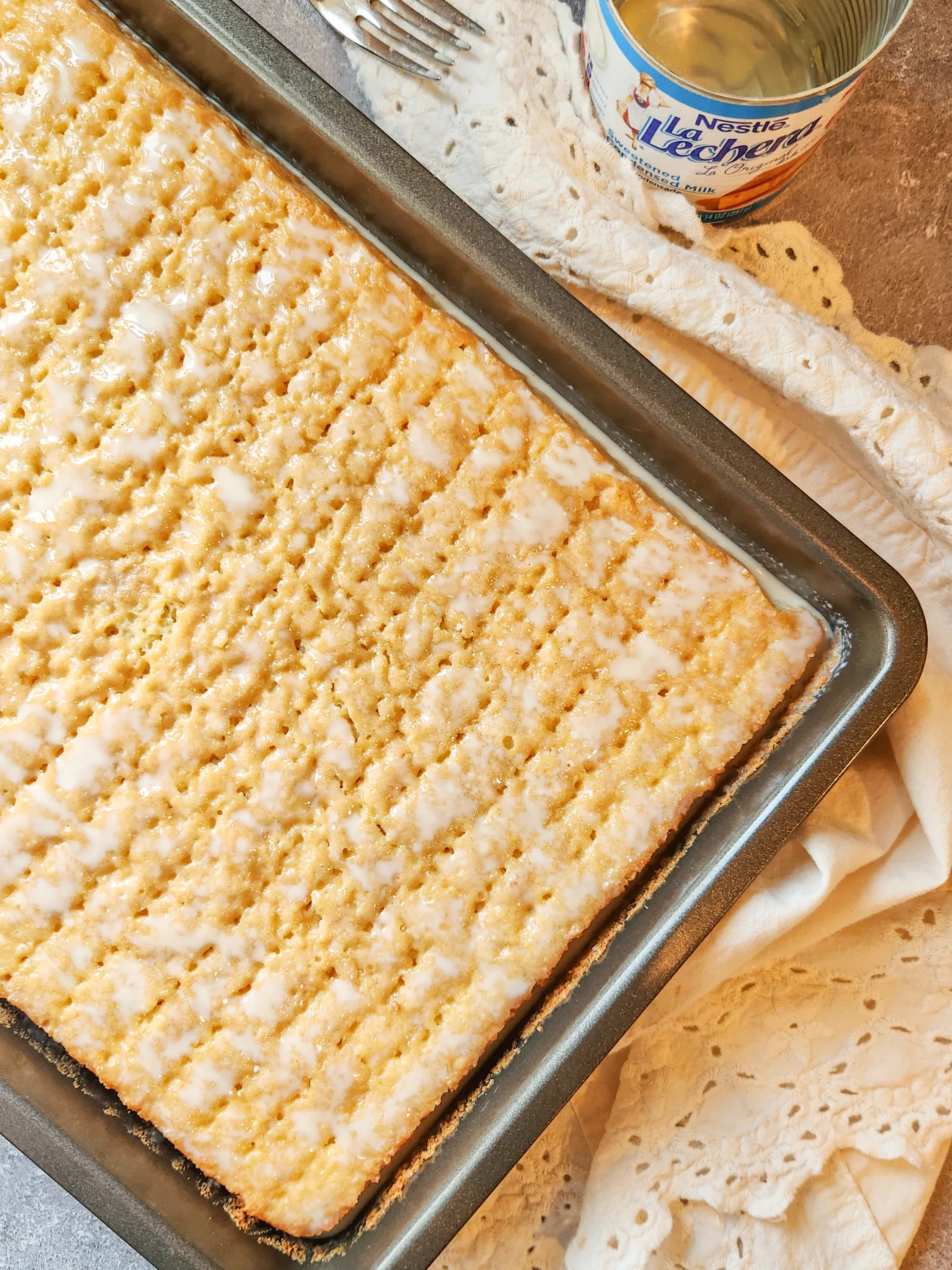 a yellow sponge cake in a metal baking pan is poked with holes and covered in  a milky mixture. It sits on a marble background with a white lace linen.