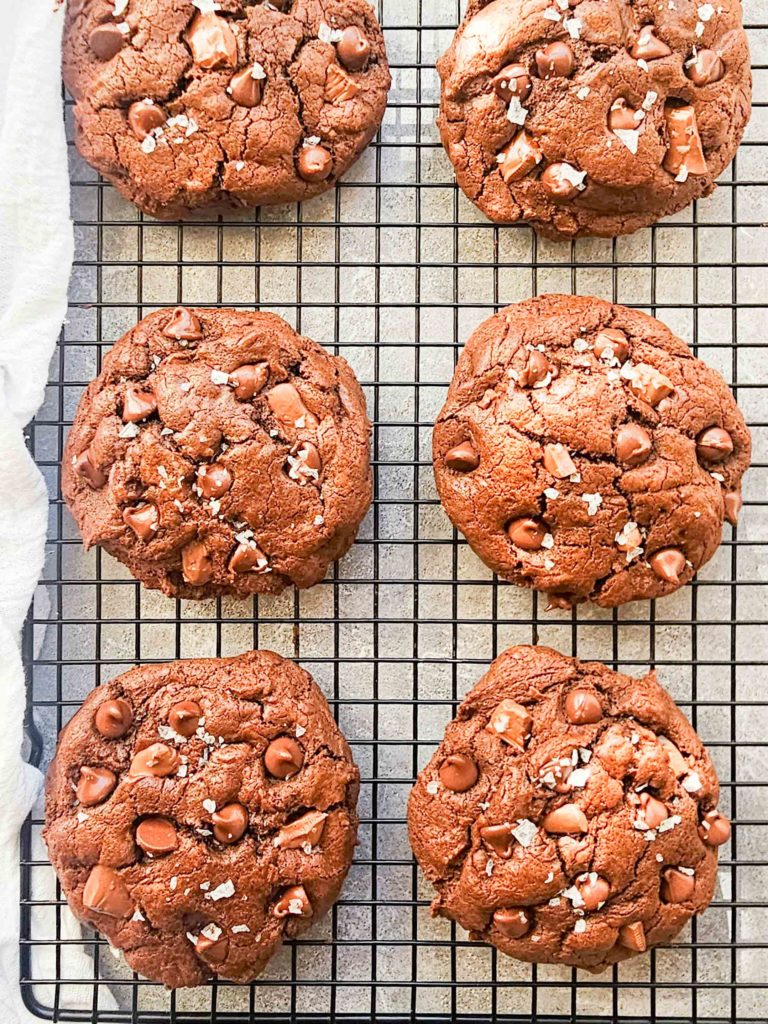 6 big and rounded chocolate cookies cool on a rack. They have lots of chocolate chips and are sprinkled with flaky salt.