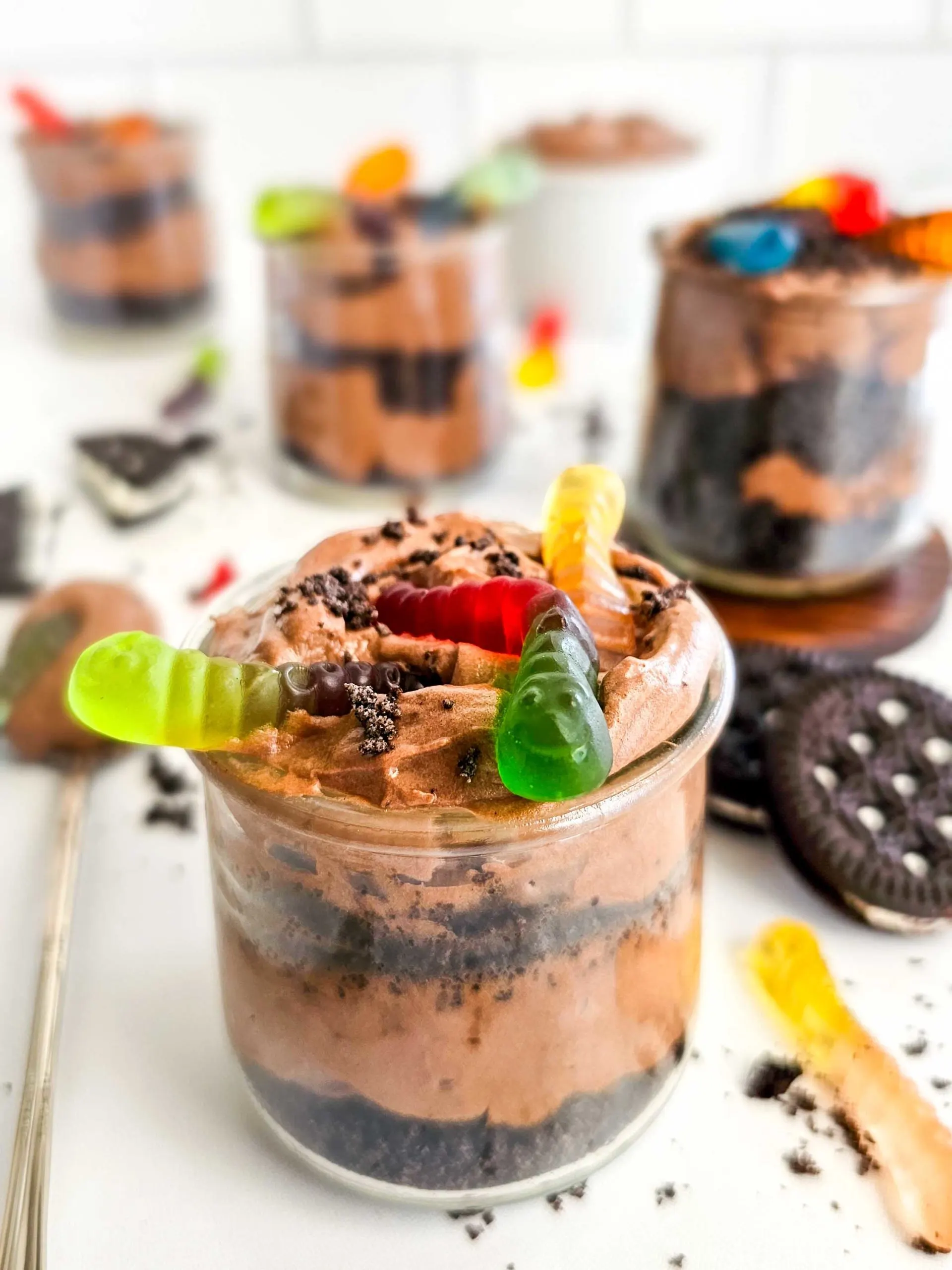 Chocolate pudding and Oreo cookie crumbs are layered in a small glass cup and topped with 3 colored gummy worms.