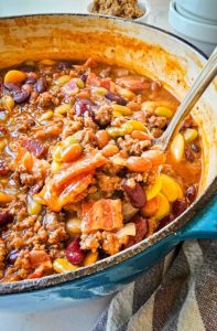Thick and hearty baked beans are shown in a blue ceramic dutch oven. A ladle is scooping some up and you can see different kinds of beans, strips of bacon, and hamburger